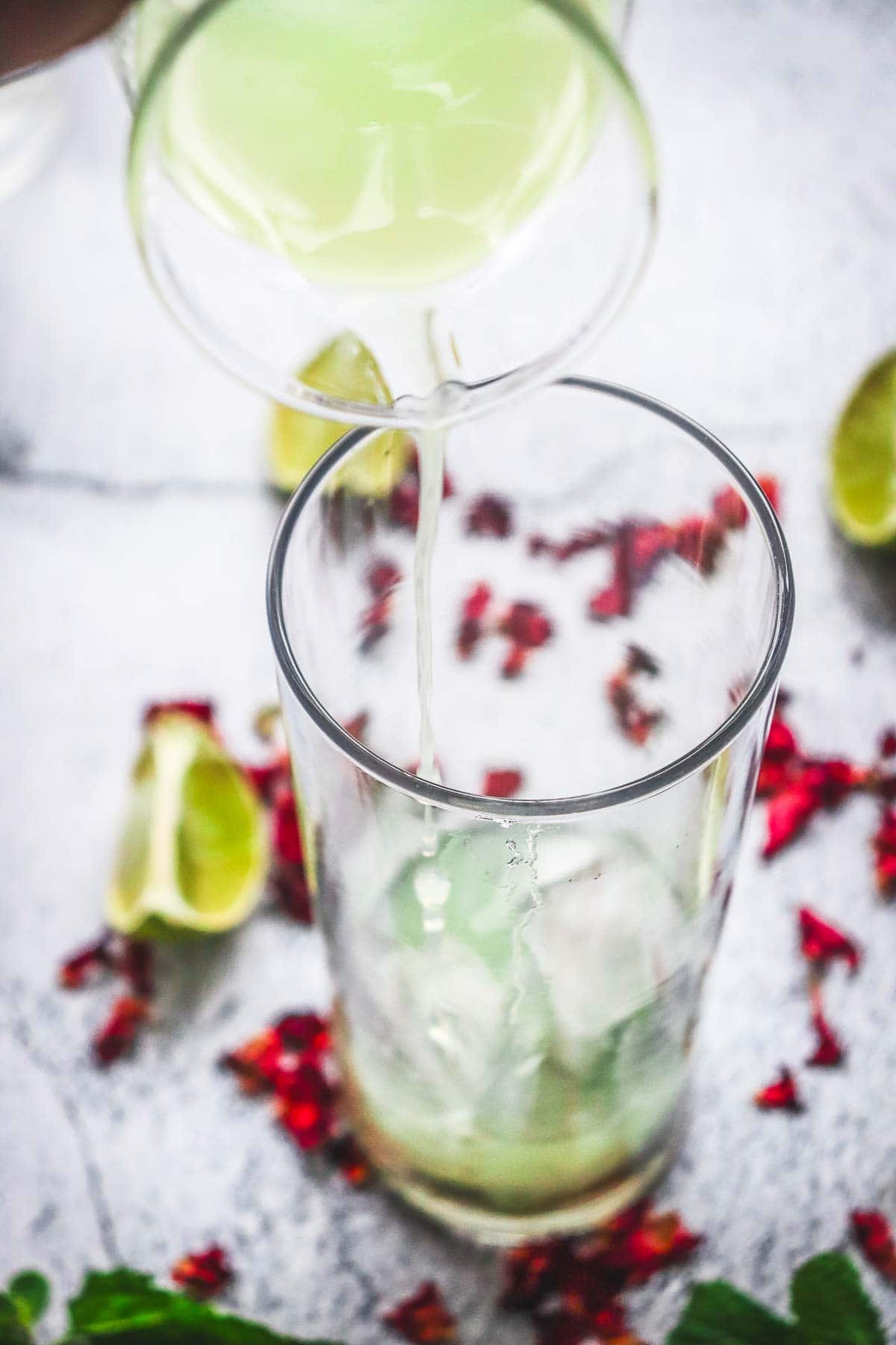 Lime juice being poured into a glass with ice, surrounded by lime wedges and red chili flakes, for a zesty non-alcoholic rose mojito mocktail recipe.