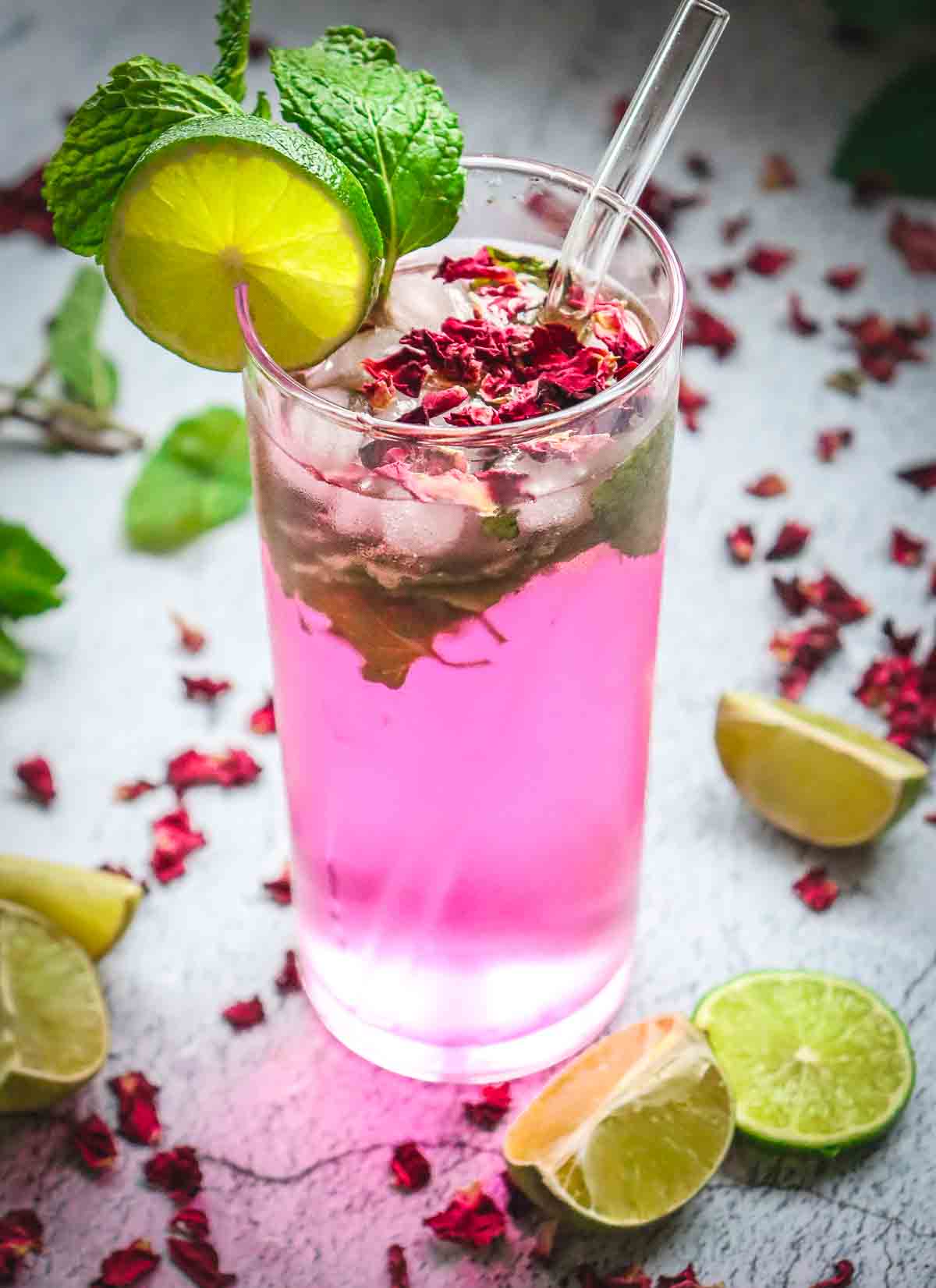 A non-alcoholic rose mojito mocktail recipe served in a tall glass with ice, mint, lime slice, and petals, topped with a straw and surrounded by fresh limes.