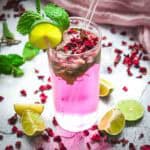 A tall glass of non-alcoholic rose mojito mocktail with ice, mint, lime slice, and rose petals, surrounded by lime wedges.