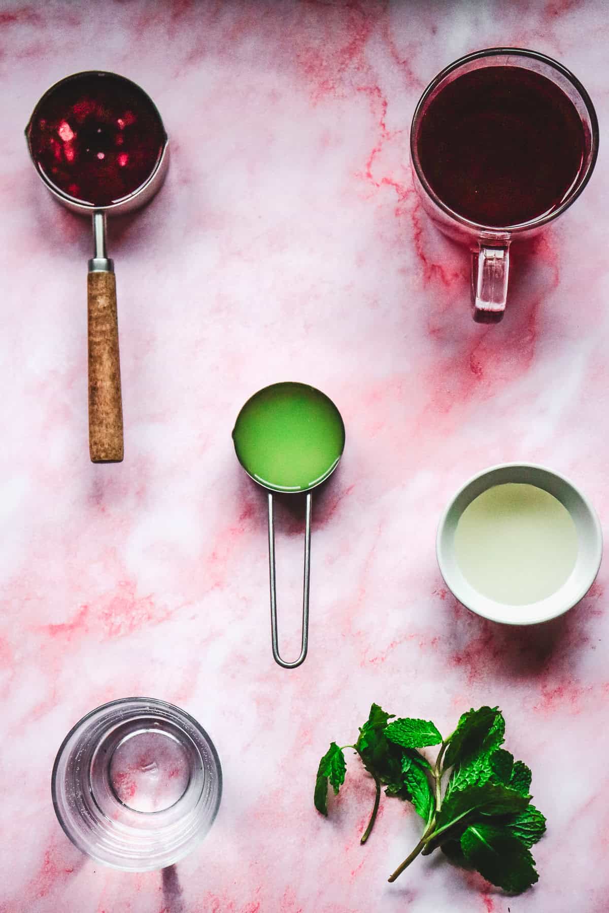 Flat lay of ingredients for a love potion Valentine's Day mocktail: pomegranate juice, lime juice, cream, water, and fresh mint on a pink marble surface.