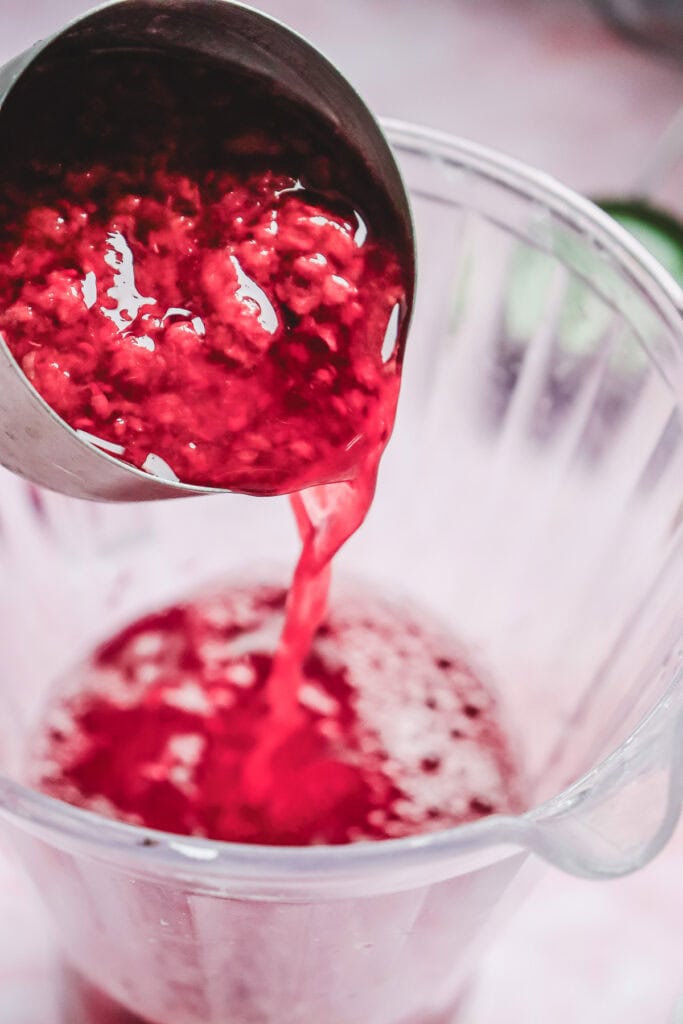 Red berry mixture, perfect for a Valentine's Day mocktail, is being poured from a ladle into a clear blender pitcher.