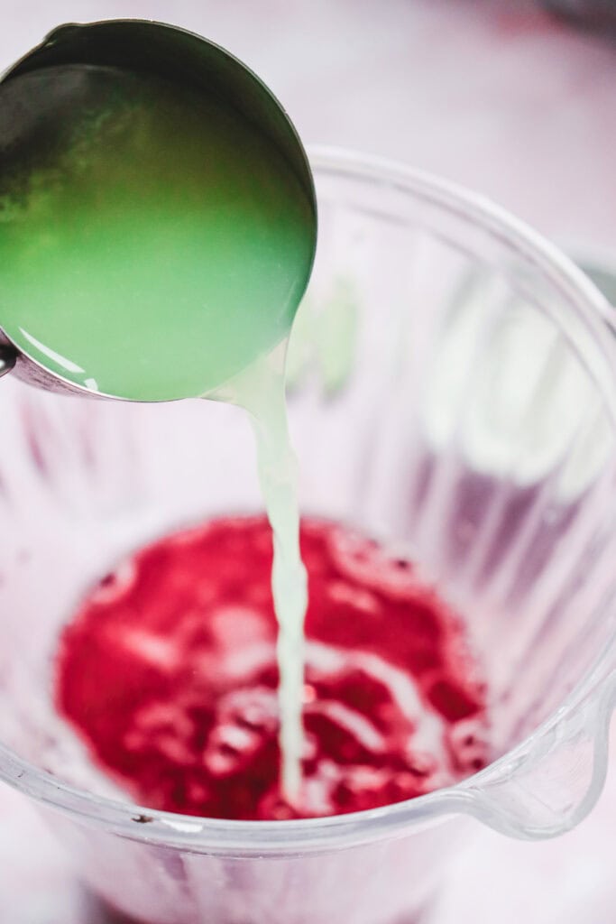 Lime juice is poured from a measuring cup into a blender with a red liquid inside, creating a festive Valentine&rsquo;s Day mocktail.
