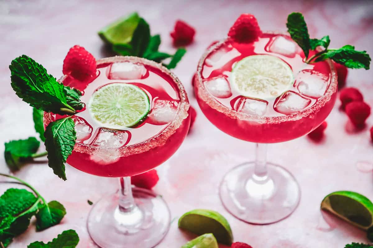 Two pink love potion mocktails with ice, lime slices, mint, and raspberries in sugar-rimmed glasses on a pink surface&mdash;perfect for Valentine&rsquo;s Day.