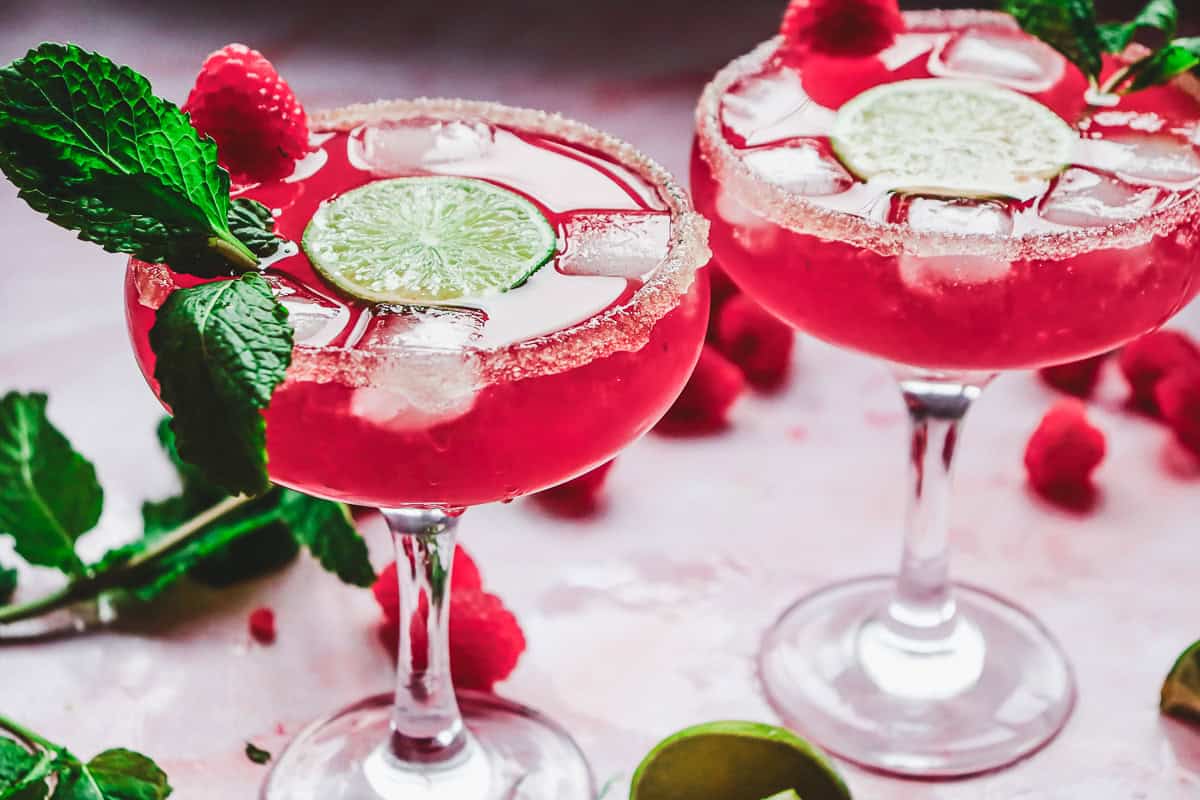 Two pink Valentine's Day mocktails in glasses with ice, raspberries, lime slices, and mint sprigs sit on a pink surface&mdash;perfect for serving as a love potion-inspired drink.
