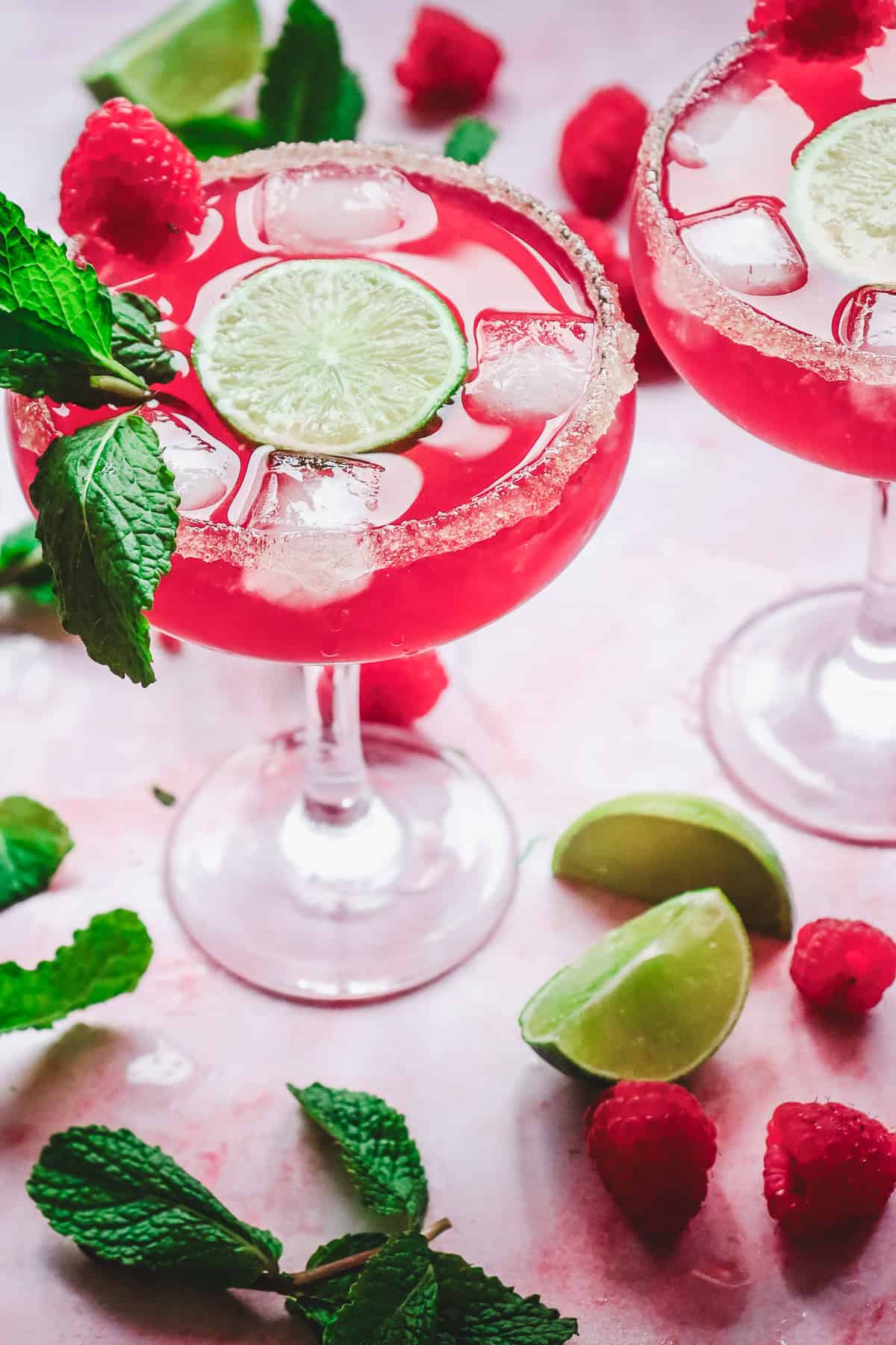 Two pink love potion mocktails with ice, lime slices, mint, and raspberries, surrounded by fresh mint and lime wedges&mdash;perfect for Valentine&rsquo;s Day.