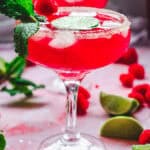 A vibrant pink Valentine's Day mocktail with ice, mint, raspberries, and lime slices in a glass, surrounded by lime wedges.