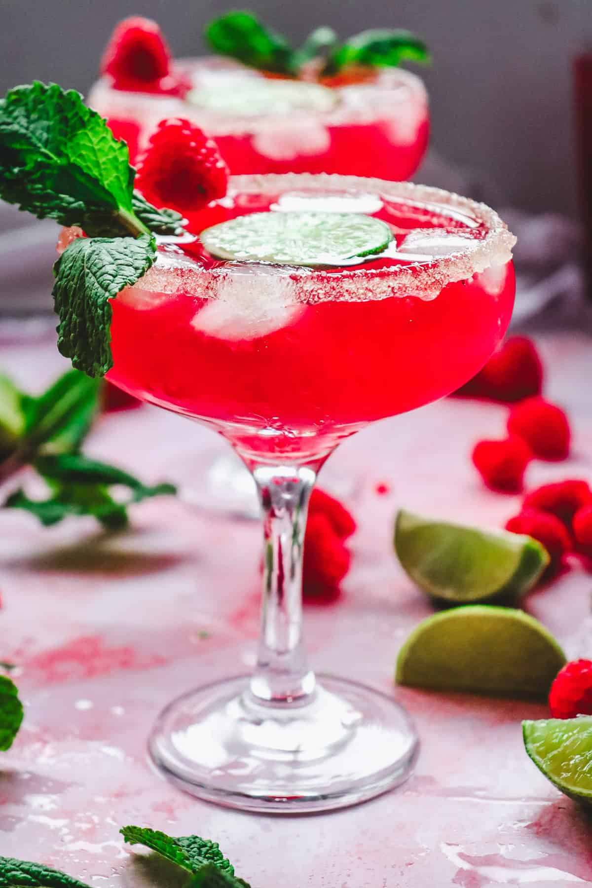 A vibrant pink Valentine's Day mocktail with ice, mint, raspberries, and lime slices in a glass, surrounded by lime wedges.