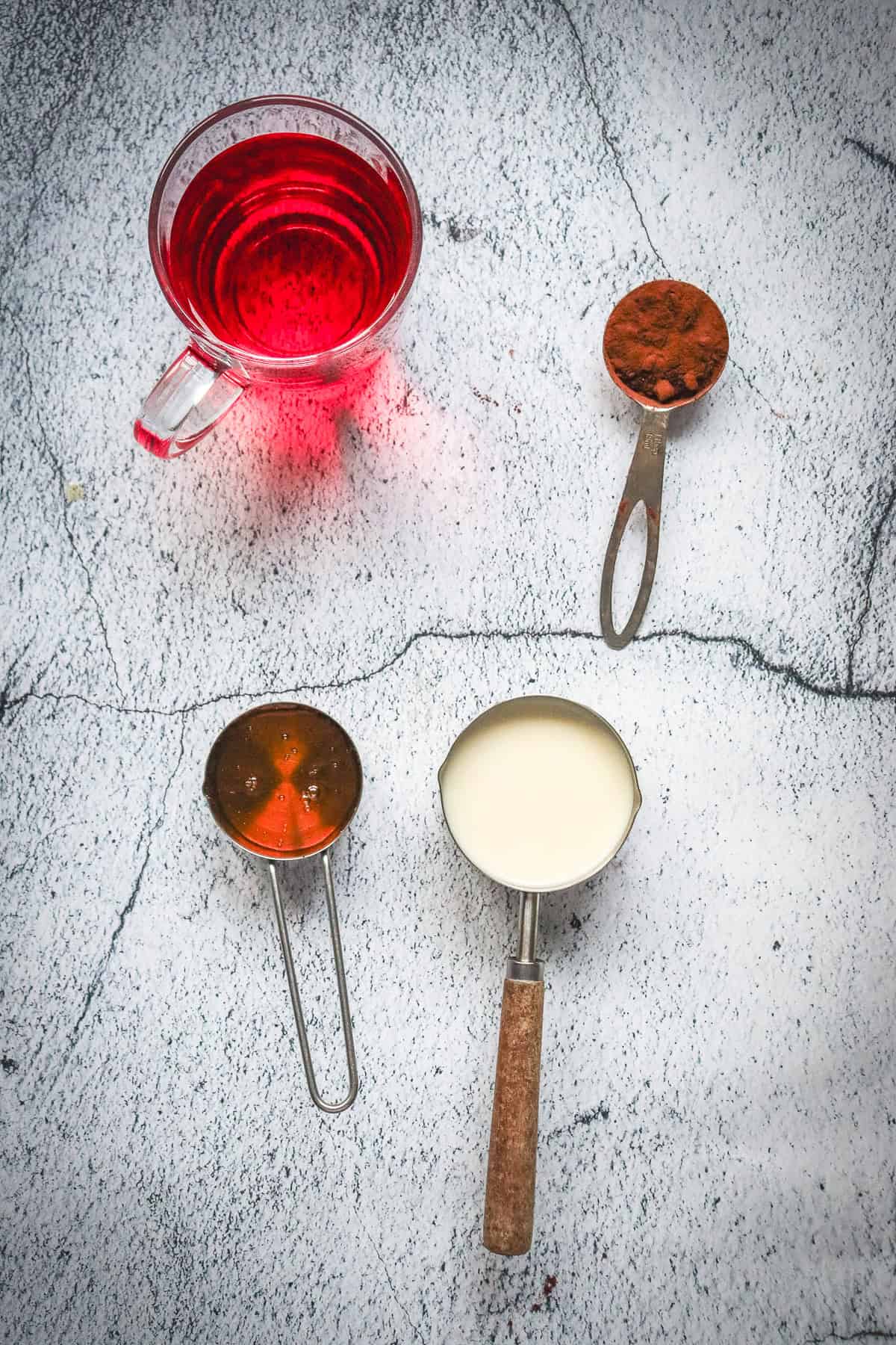 Glass cup of red liquid, inspired by a Red Velvet Mocktail Recipe, with three measuring cups holding cocoa powder, honey, and milk on a gray textured surface.