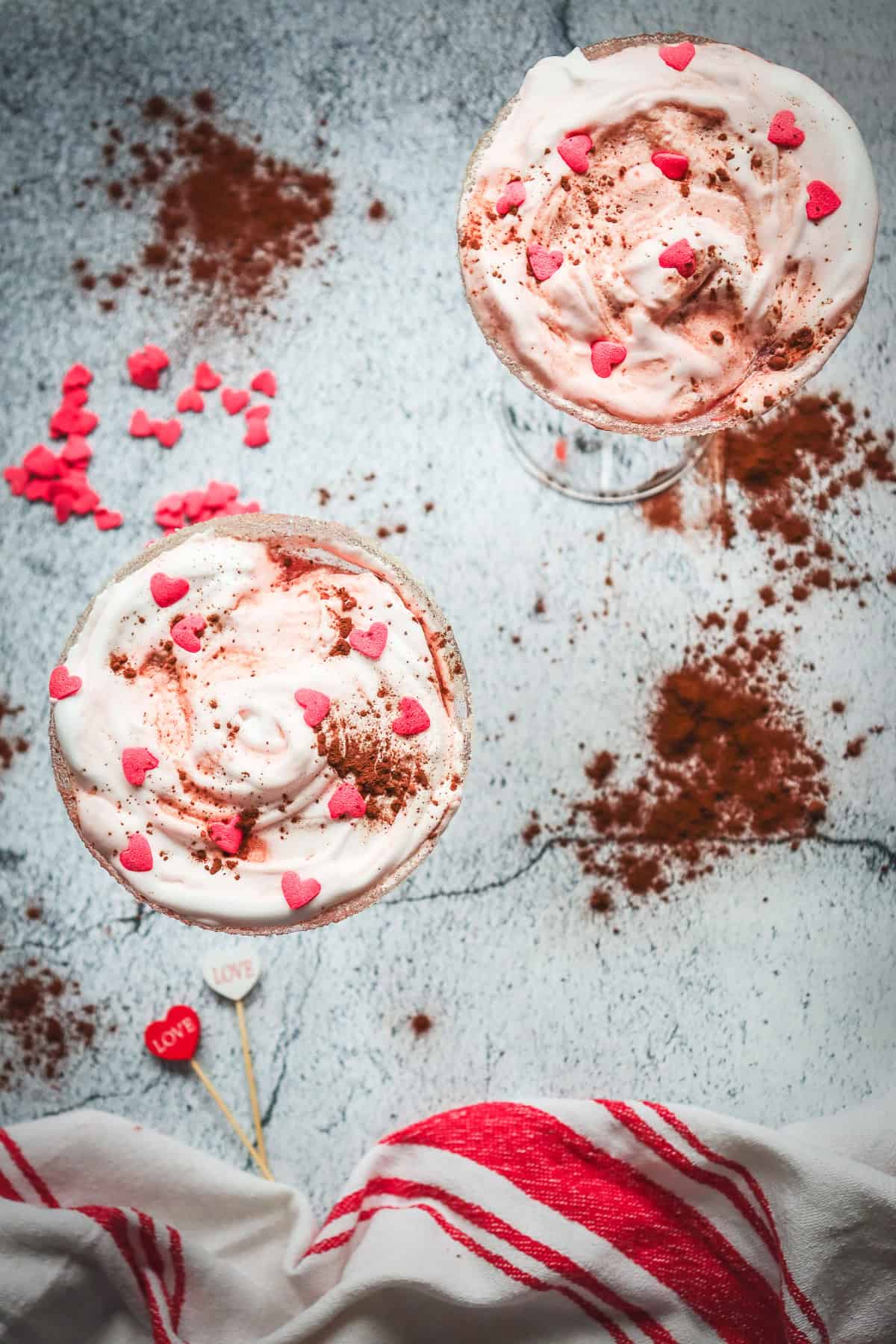 Two creamy pink Valentine mocktail desserts topped with heart sprinkles and cocoa sit on a red-striped cloth, surrounded by scattered hearts.