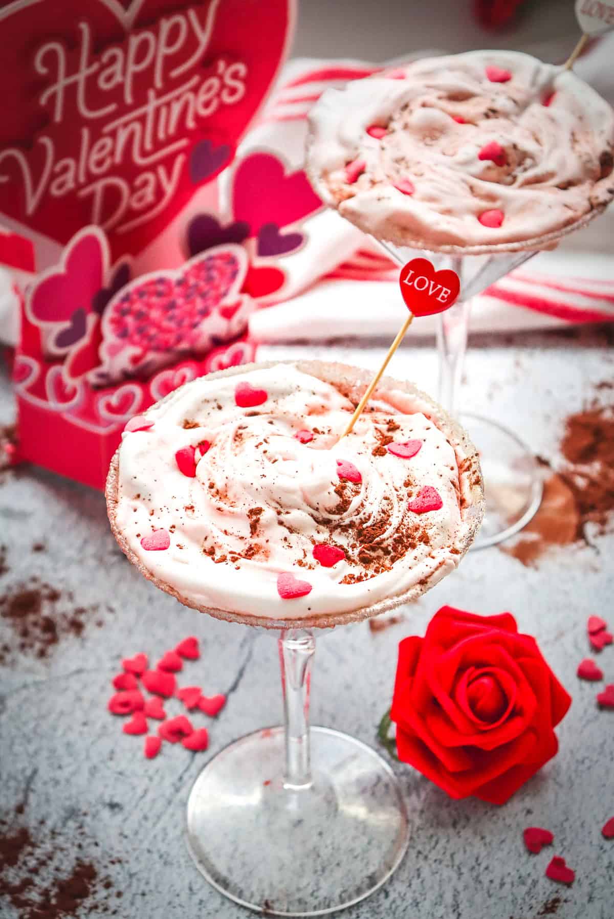 Two dessert glasses with whipped cream, heart sprinkles, a red rose, and a Valentine&rsquo;s Day card in the background&mdash;an inviting scene perfect for serving your favorite Red Velvet mocktail recipe.