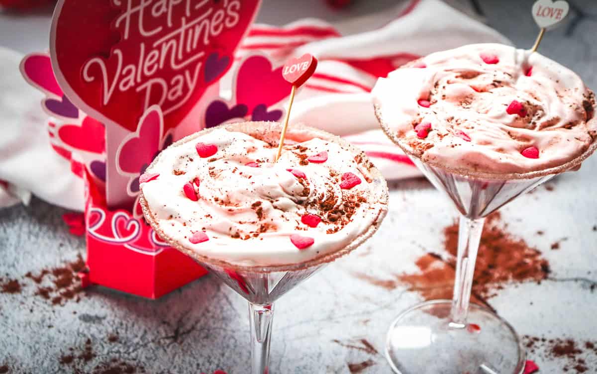 Two pink Valentine&rsquo;s drinks in martini glasses with heart sprinkles and &ldquo;Love&rdquo; toppers, festive card in background&mdash;a delicious Chocolate Martini Mocktail that&rsquo;s the perfect Valentine recipe.