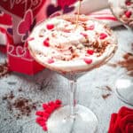 A creamy Red Velvet Valentine's Day dessert in a glass, topped with heart sprinkles and a "love" pick, sits beside a red rose.