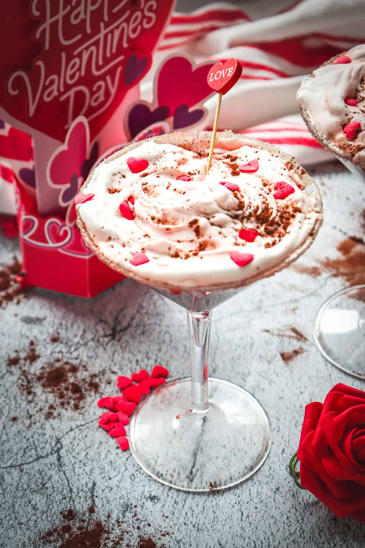 A creamy Red Velvet Valentine's Day dessert in a glass, topped with heart sprinkles and a "love" pick, sits beside a red rose.