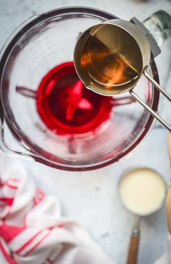 A measuring cup pours liquid into a blender with red liquid for a delicious Red Velvet Martini, next to a white cup and striped towel.