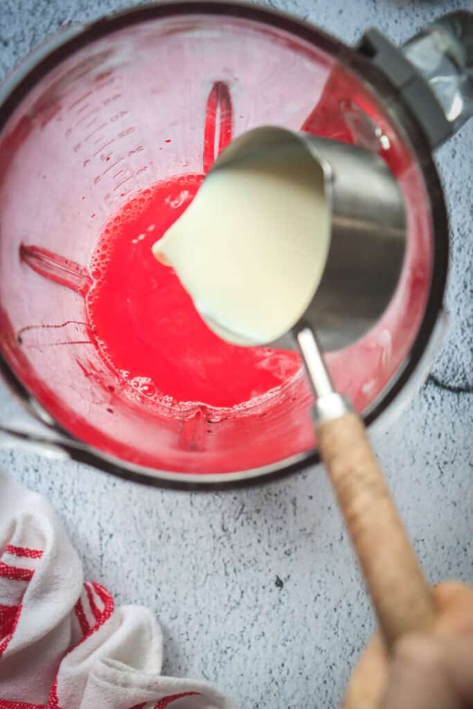 A metal cup pours milk into a bowl with a red liquid mixture on a light surface, creating the perfect base for a Red Velvet mocktail recipe.