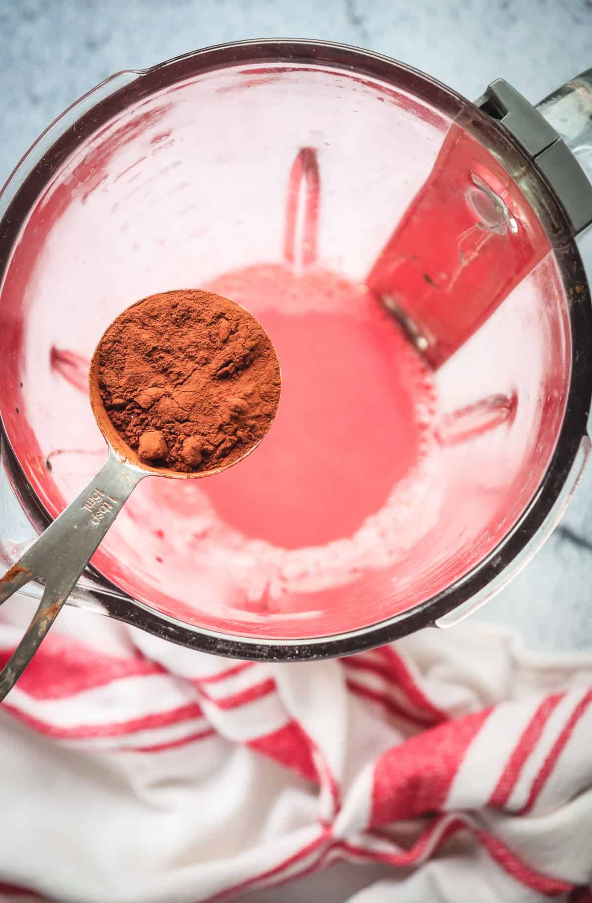 A measuring spoon of cocoa powder held over a blender with pink liquid inside, perfect for creating a festive Red Velvet or Valentine-inspired Chocolate Martini, sits beside a red-striped towel.