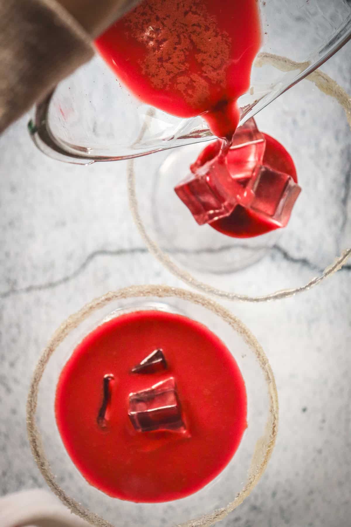 Red Velvet Martini being poured over ice into a glass, next to another of the same vibrant drink, both resting on a marble surface.