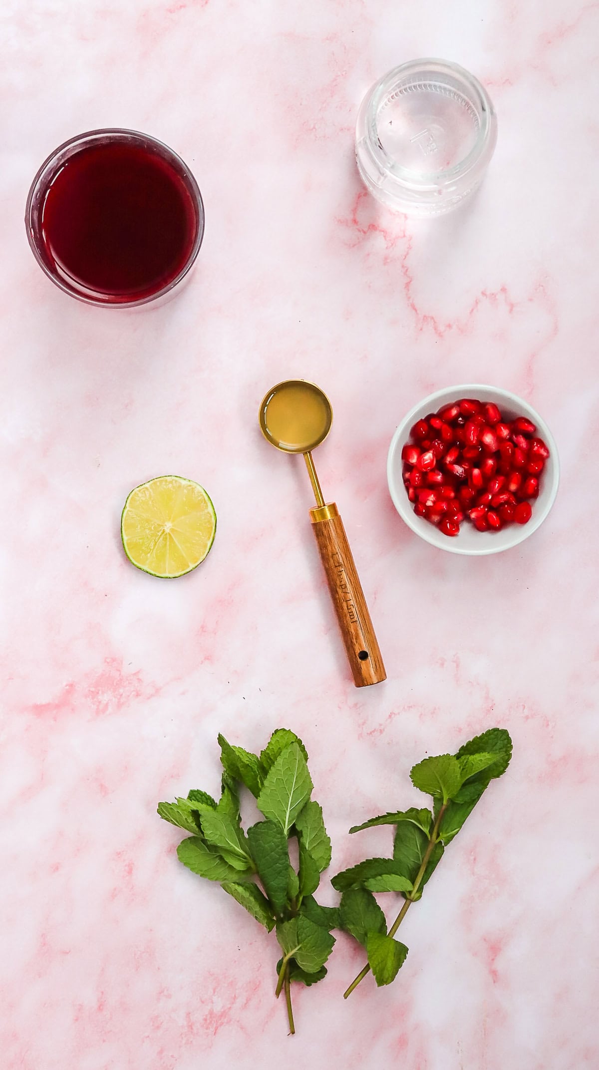 Ingredients on a pink surface: pomegranate juice, water, honey, lime, pomegranate seeds, and mint leaves&mdash;perfect for crafting a refreshing Pomegranate Mocktail.