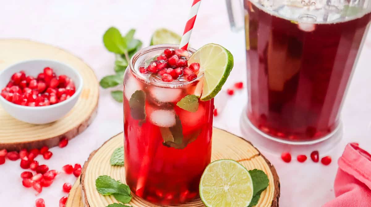 A glass of Virgin Pomegranate Mocktail with mint, lime slice, and a striped straw, surrounded by pomegranate seeds.