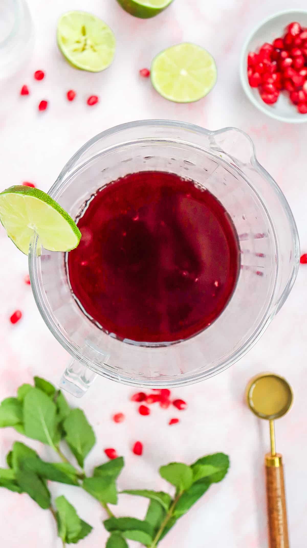 Top view of a pitcher with red Pomegranate Fizz Mocktail, garnished with a lime slice and surrounded by fresh lime, mint, and pomegranate.