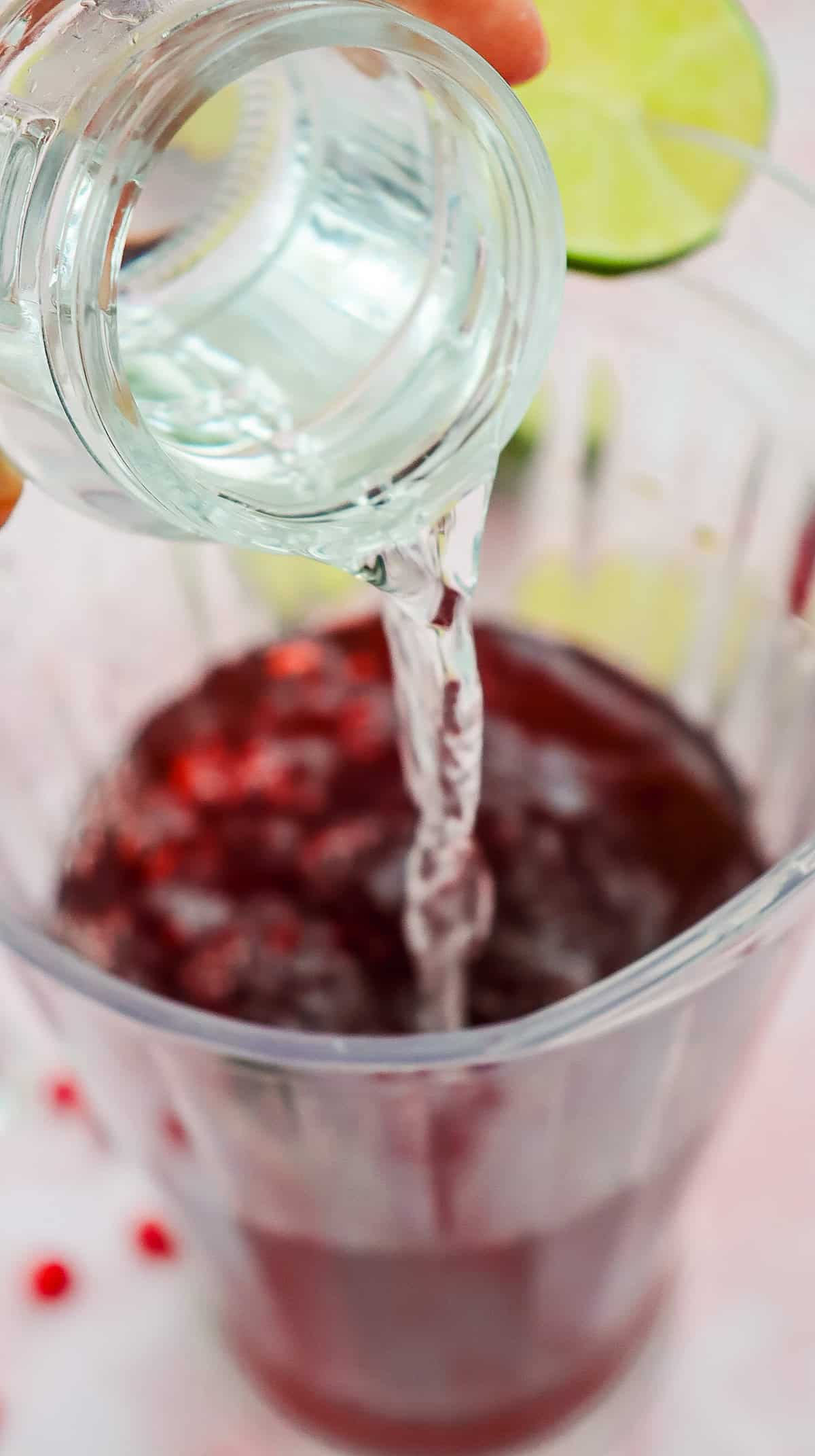A clear liquid is being poured from a small glass into a Virgin Pomegranate mocktail garnished with a lime slice on the rim.