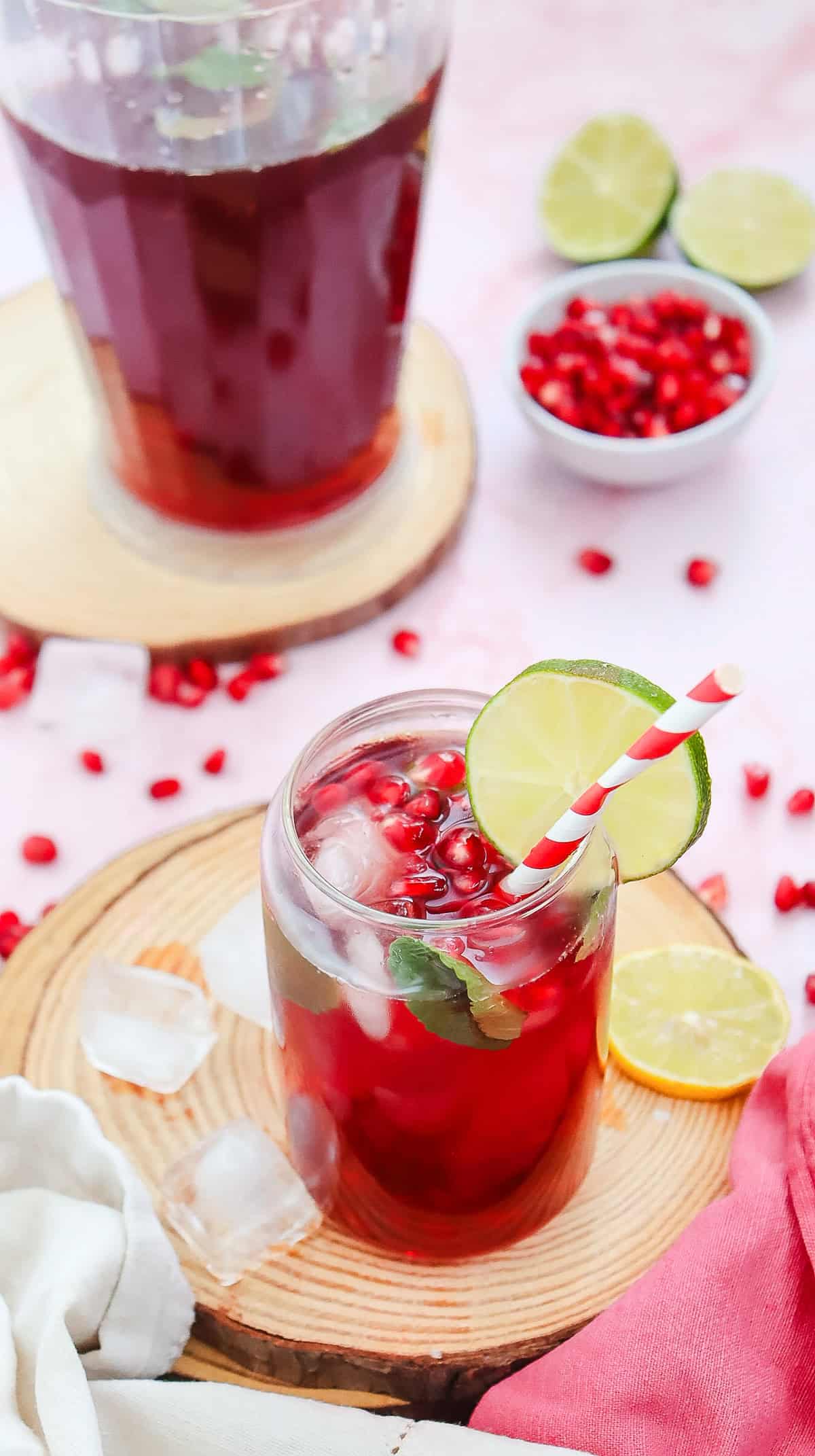 A glass of Virgin Pomegranate Mocktail with ice, lime slice, and a straw, surrounded by fresh lime and pomegranate seeds.