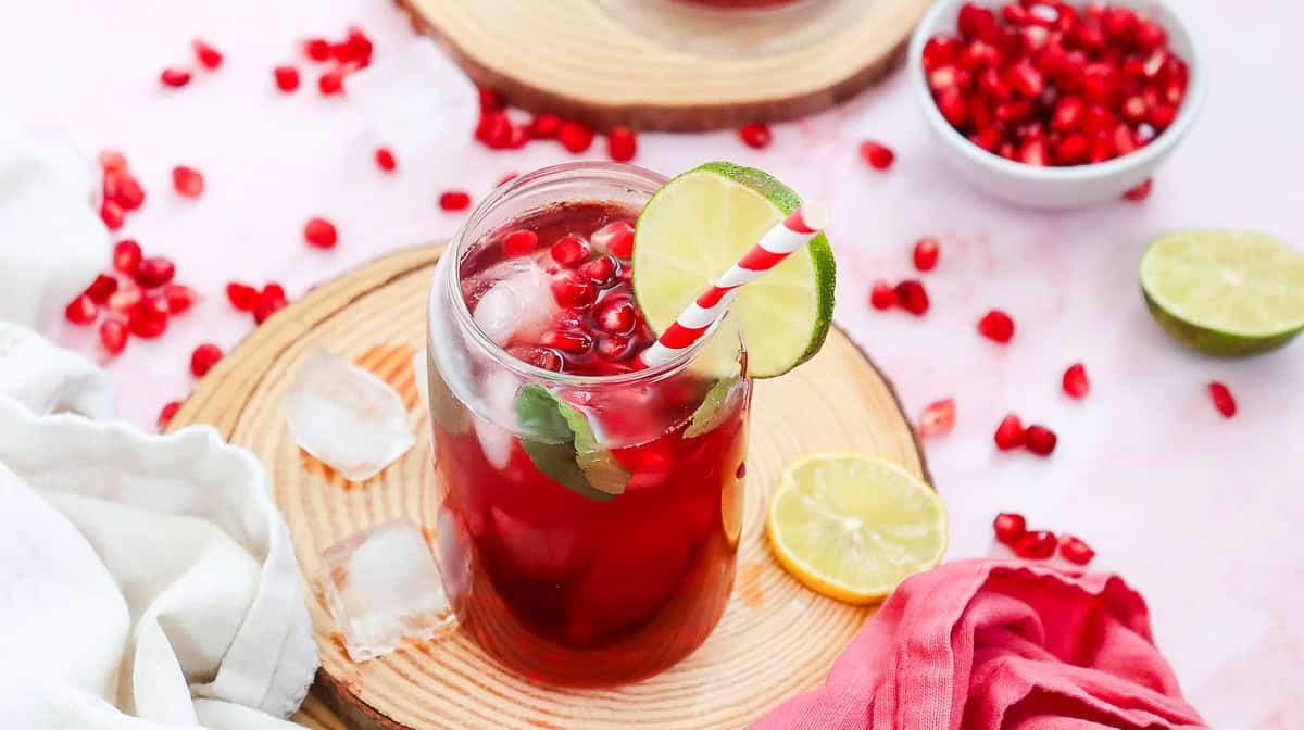 A glass of red pomegranate virgin mocktail with ice, a lime slice, and a striped straw, surrounded by fresh pomegranate seeds.