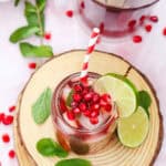 A glass of iced pomegranate Fizz Mocktail with mint, lime slices, and a red-striped straw sits on a wooden coaster.