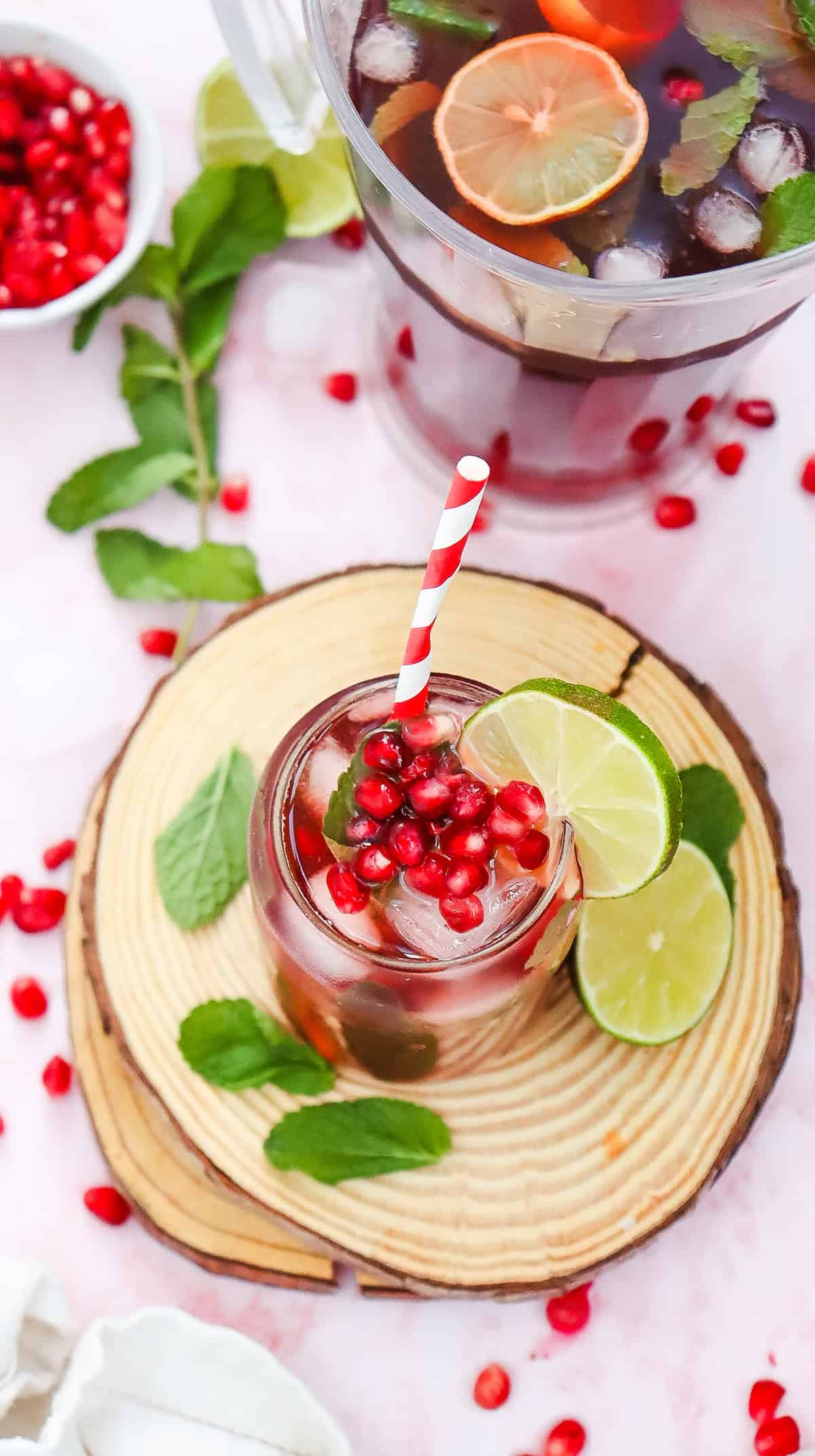 A glass of iced pomegranate Fizz Mocktail with mint, lime slices, and a red-striped straw sits on a wooden coaster.