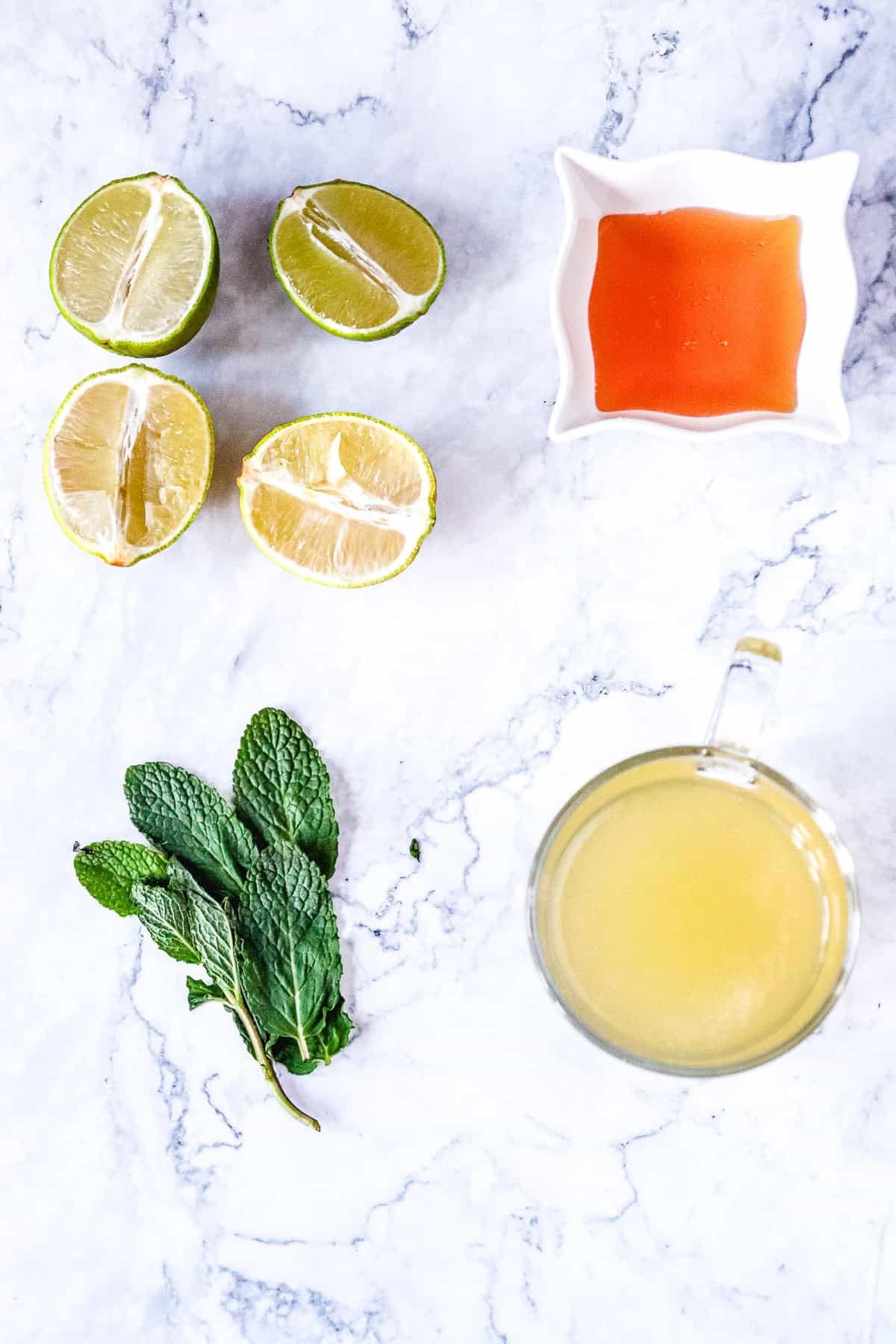 Flat lay of halved limes, honey in a dish, mint leaves, and a mug of yellow liquid on marble&mdash;perfect ingredients for crafting a refreshing Virgin Mojito or trying out a classic mocktail recipe at home.