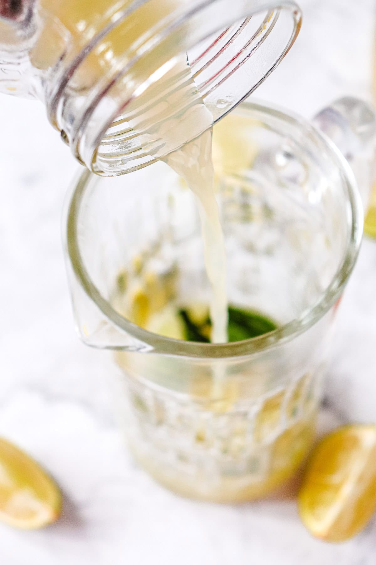 Liquid is being poured from a jar into a glass measuring cup with lime wedges and juice on a white surface, perfect for preparing a refreshing Virgin Mojito or Classic Mocktail.