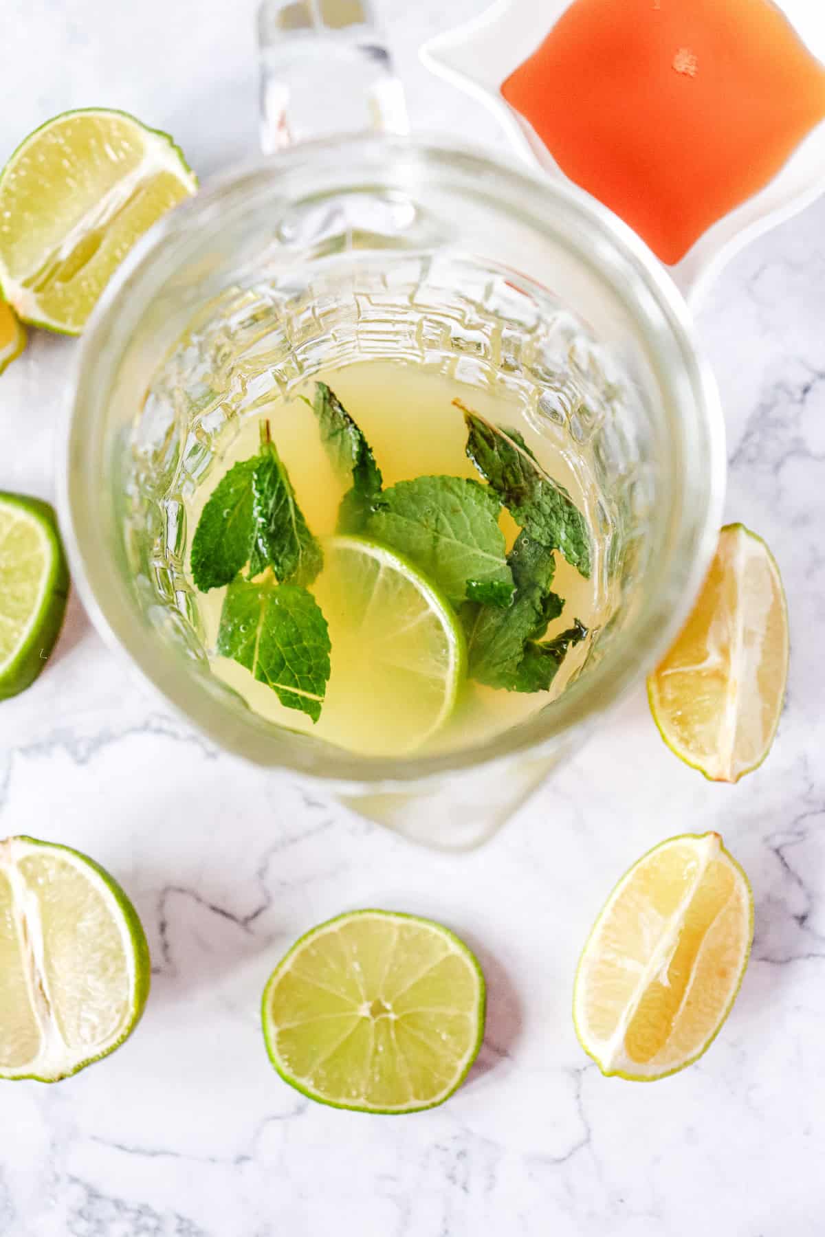 A glass jug with lime slices, mint leaves, and juice, surrounded by cut limes and a dish of red sauce&mdash;perfect for serving a refreshing Virgin Mojito mocktail recipe.