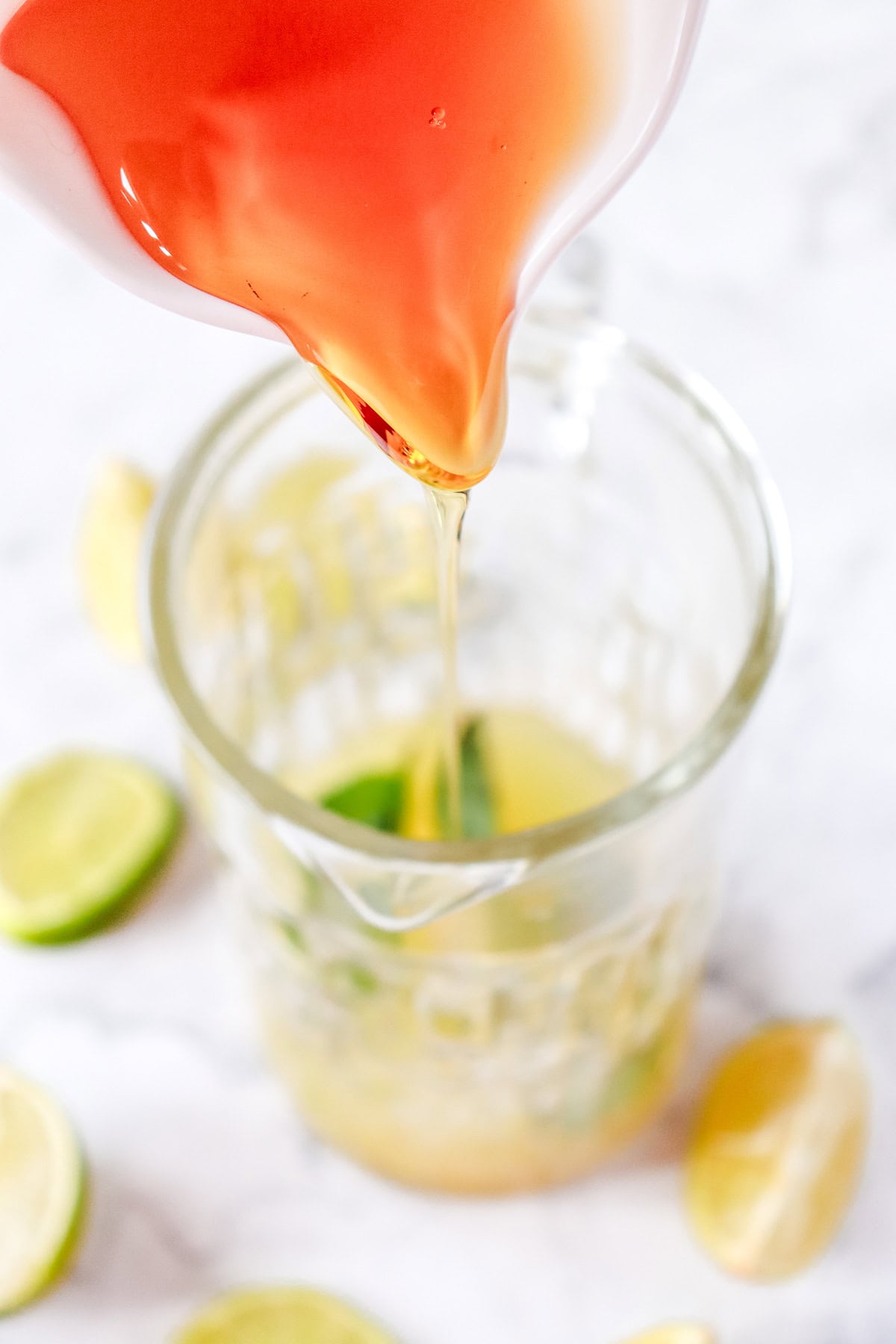 Red liquid being poured into a glass pitcher with lime slices and juice on a white surface, perfect for a refreshing Virgin Mojito or to inspire your next mocktail recipe.