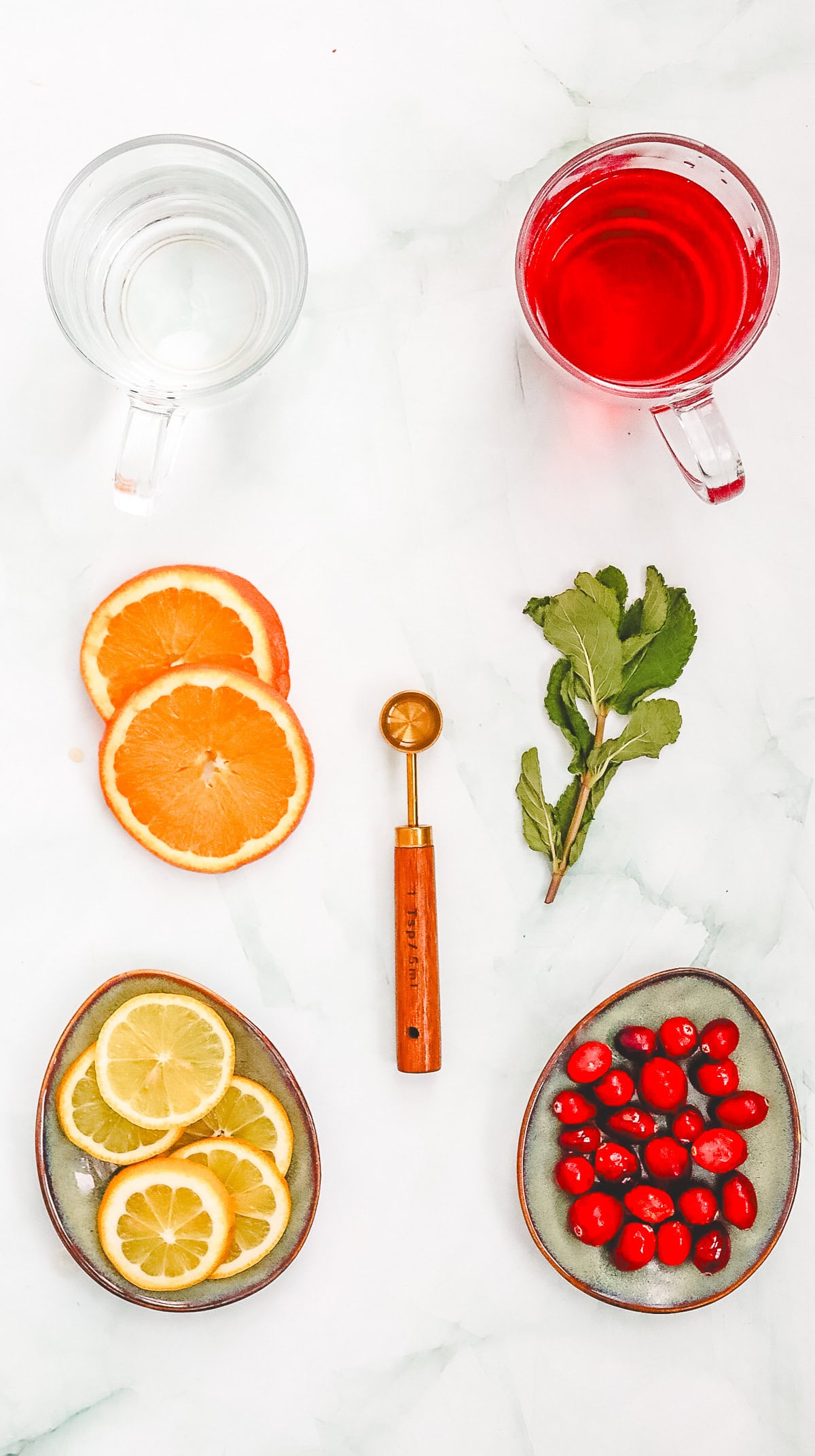Two cups of Cranberry Spritzer, a refreshing non-alcoholic mocktail, are filled with clear and red liquid, citrus slices, cranberries, and mint, with a melon baller resting on the marble surface.