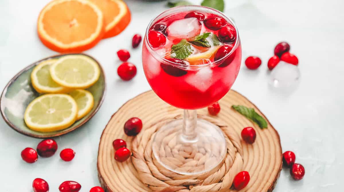 A glass of cranberry spritzer mocktail with ice, mint, and orange slice, surrounded by cranberries and citrus slices.