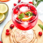 A glass of refreshing Cranberry Spritzer mocktail with ice, cranberries, orange slice, and mint on a wooden coaster.