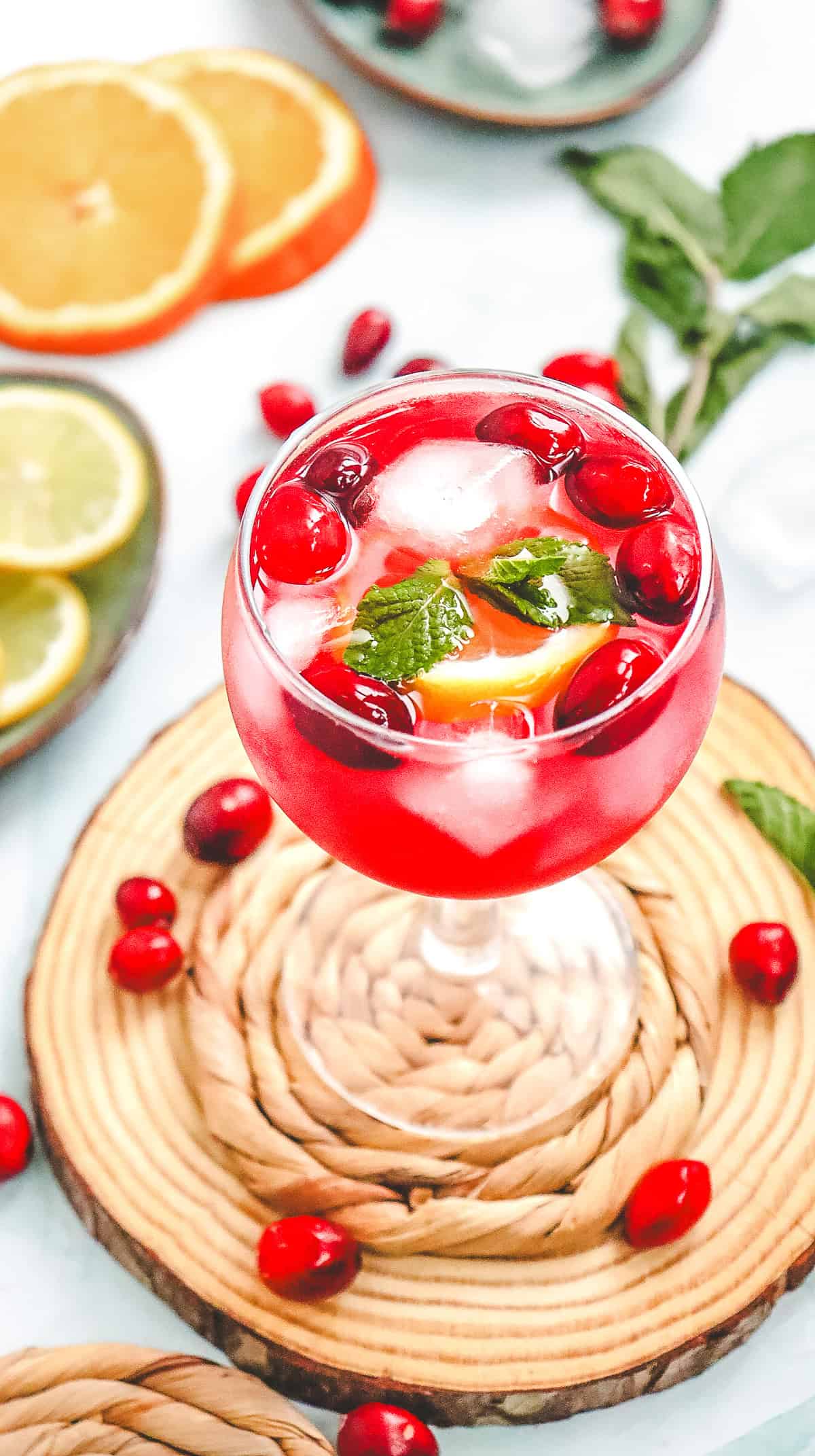 A glass of refreshing Cranberry Spritzer mocktail with ice, cranberries, orange slice, and mint on a wooden coaster.