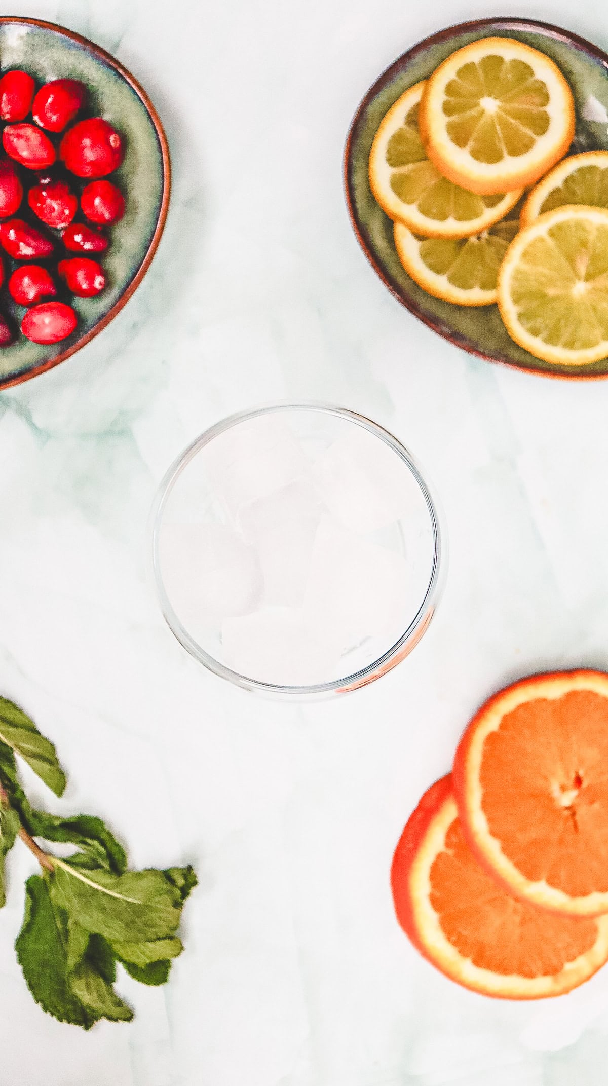 A glass of ice is surrounded by bowls of cranberries, citrus slices, and fresh mint on a white surface&mdash;perfect for crafting a refreshing Cranberry Spritzer mocktail.
