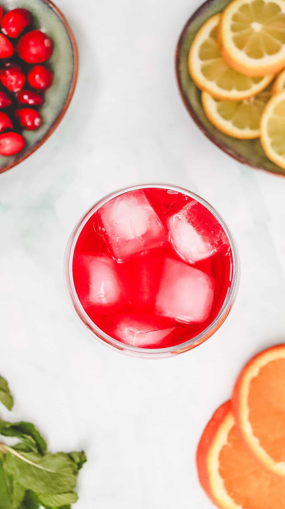 A glass of red iced Cranberry Spritzer mocktail, surrounded by cranberries, lemon and orange slices, and fresh mint leaves. This refreshing non-alcoholic drink is perfect for any occasion.