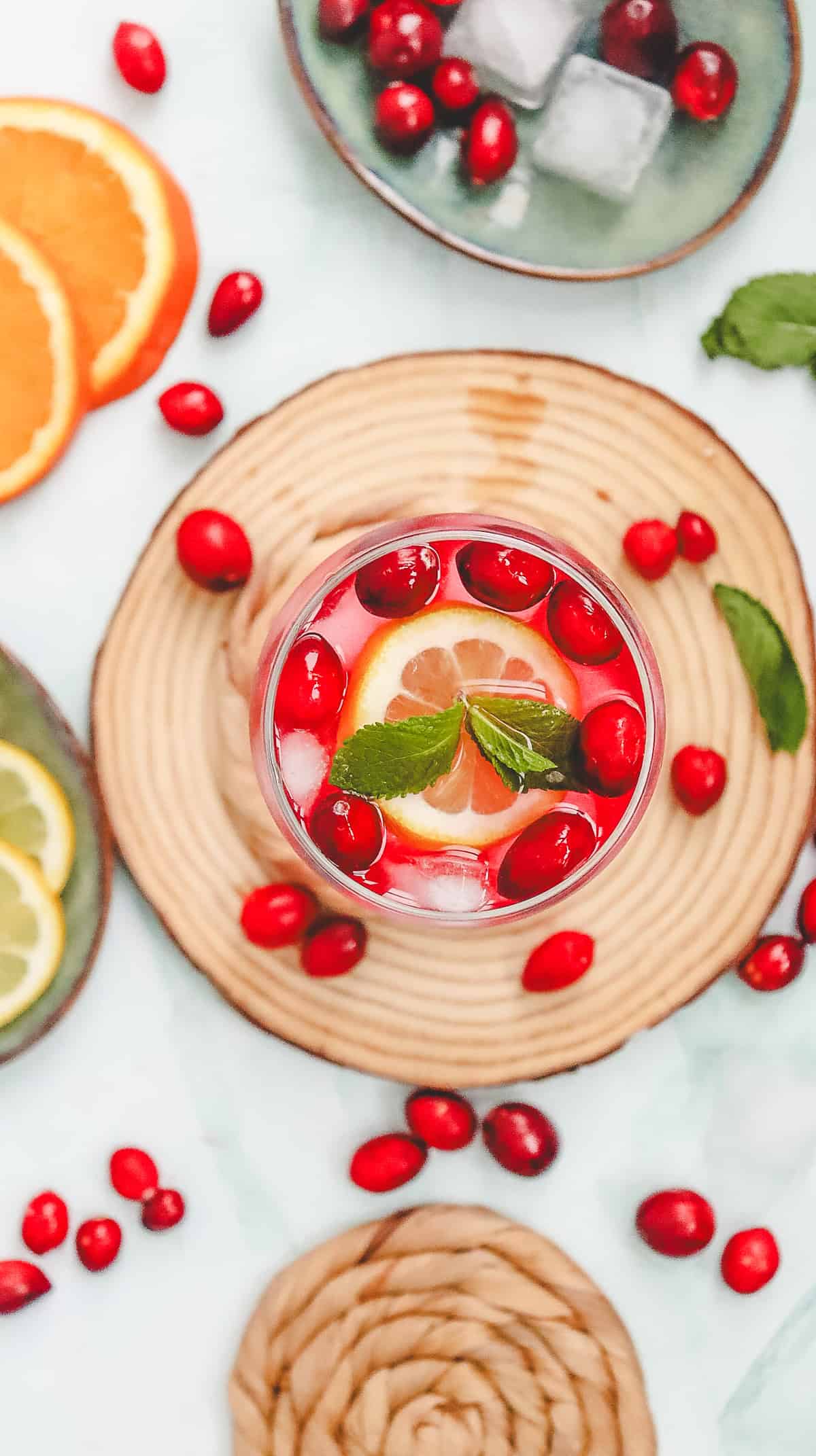 A glass of cranberry spritzer mocktail with lemon, mint, and ice, surrounded by fresh cranberries and citrus slices.