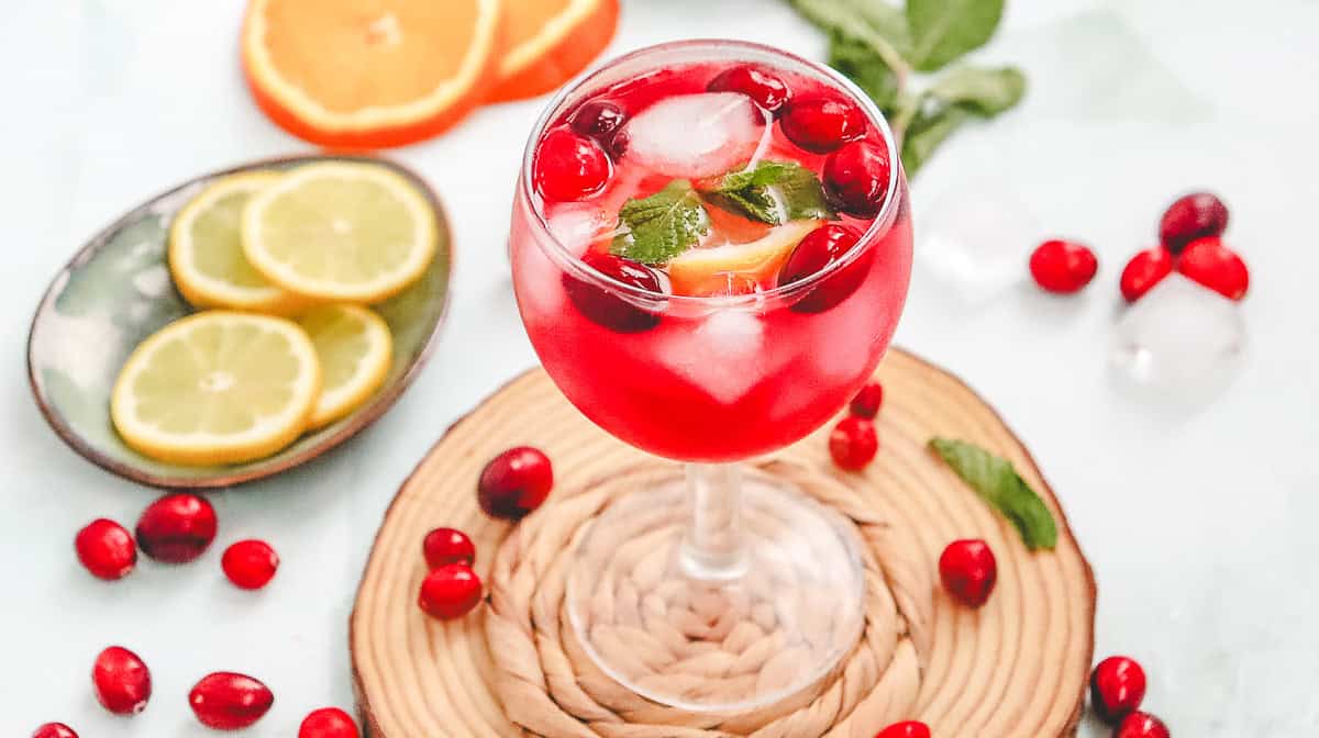 A glass of Cranberry Spritzer mocktail with ice, lemon, cranberries, and mint, surrounded by sliced citrus and cranberries.