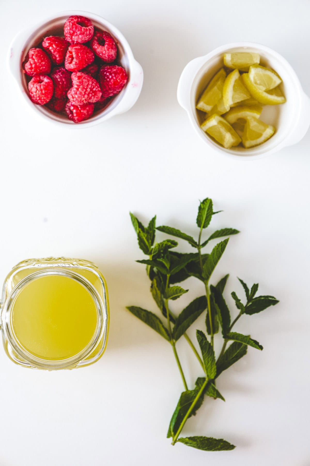 A jar of yellow liquid, fresh mint sprigs, raspberries, and lemon wedges on a white surface&mdash;perfect ingredients for a refreshing raspberry mojito mocktail recipe.