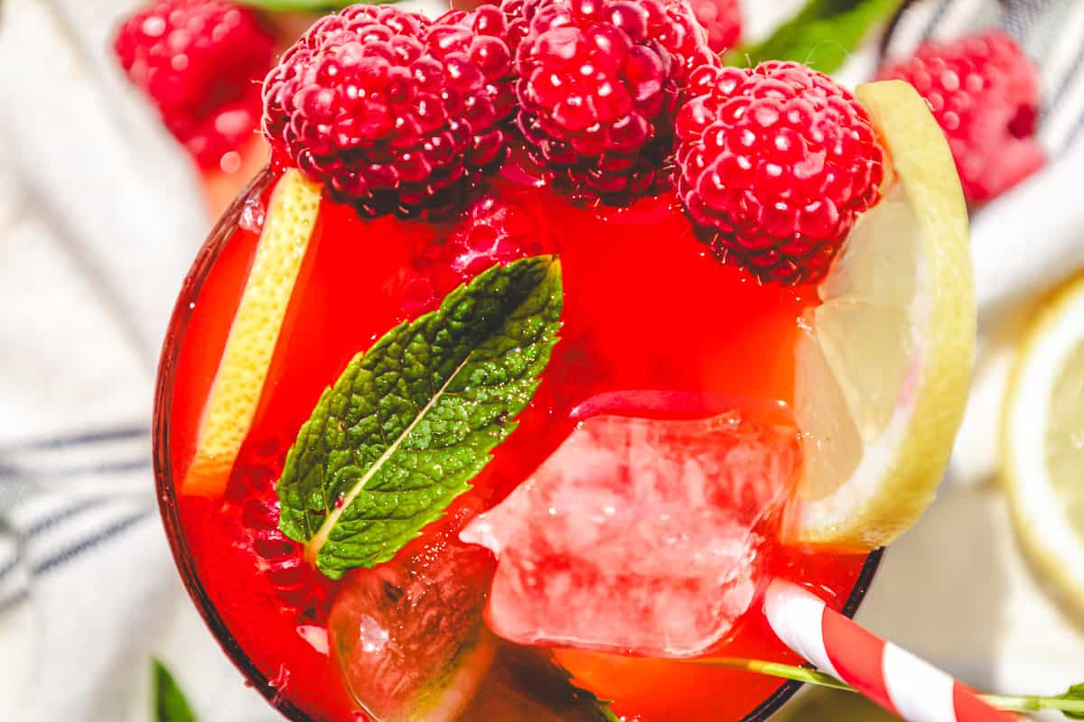 A close-up of a red raspberry mojito mocktail recipe with raspberries, lemon slices, mint leaves, ice, and a striped straw.