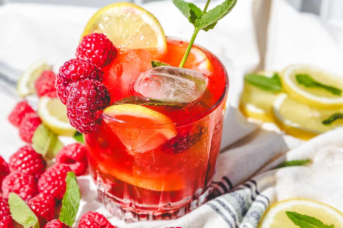 A glass of red fruit drink with ice, lemon slices, raspberries, and a mint sprig, surrounded by fresh fruit&mdash;perfect for trying a refreshing raspberry mojito mocktail recipe.