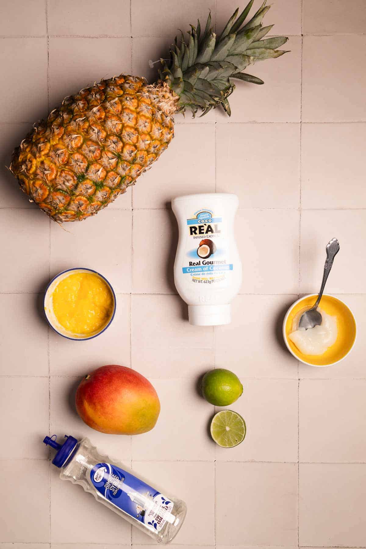 Pineapple, mango, lime, yogurt, bottled honey, and a small bowl of puree arranged on a tiled surface&mdash;perfect ingredients for a refreshing Mango Colada recipe.