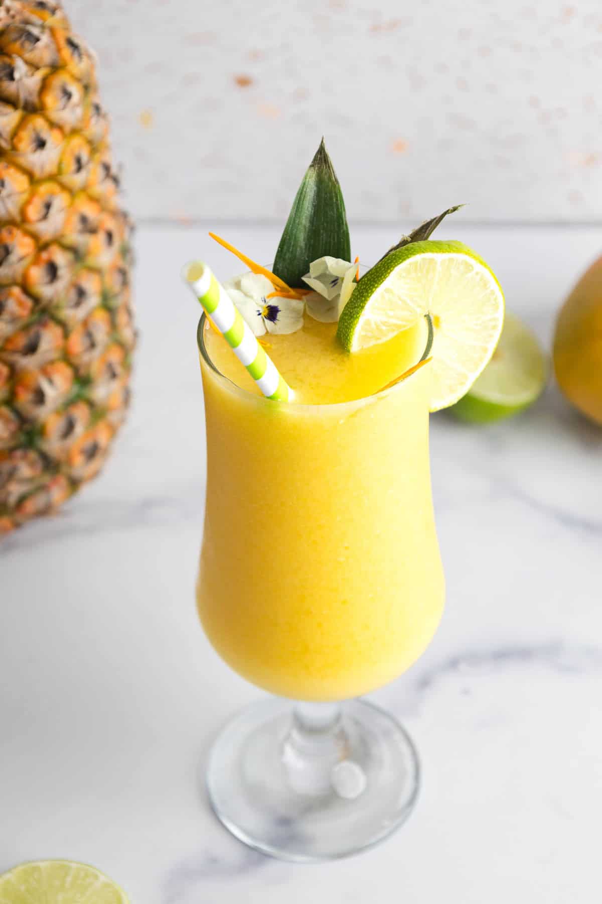 A yellow Mango Mocktail smoothie in a glass, garnished with a lime slice, flowers, and striped straws.