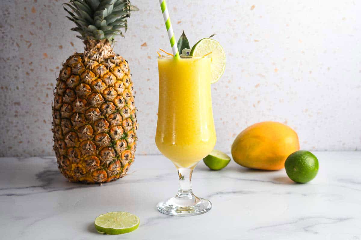 A glass of yellow smoothie with lime and straws, next to a pineapple, mango, and lime on a white surface&mdash;perfect for trying as a refreshing Mango Mocktail.