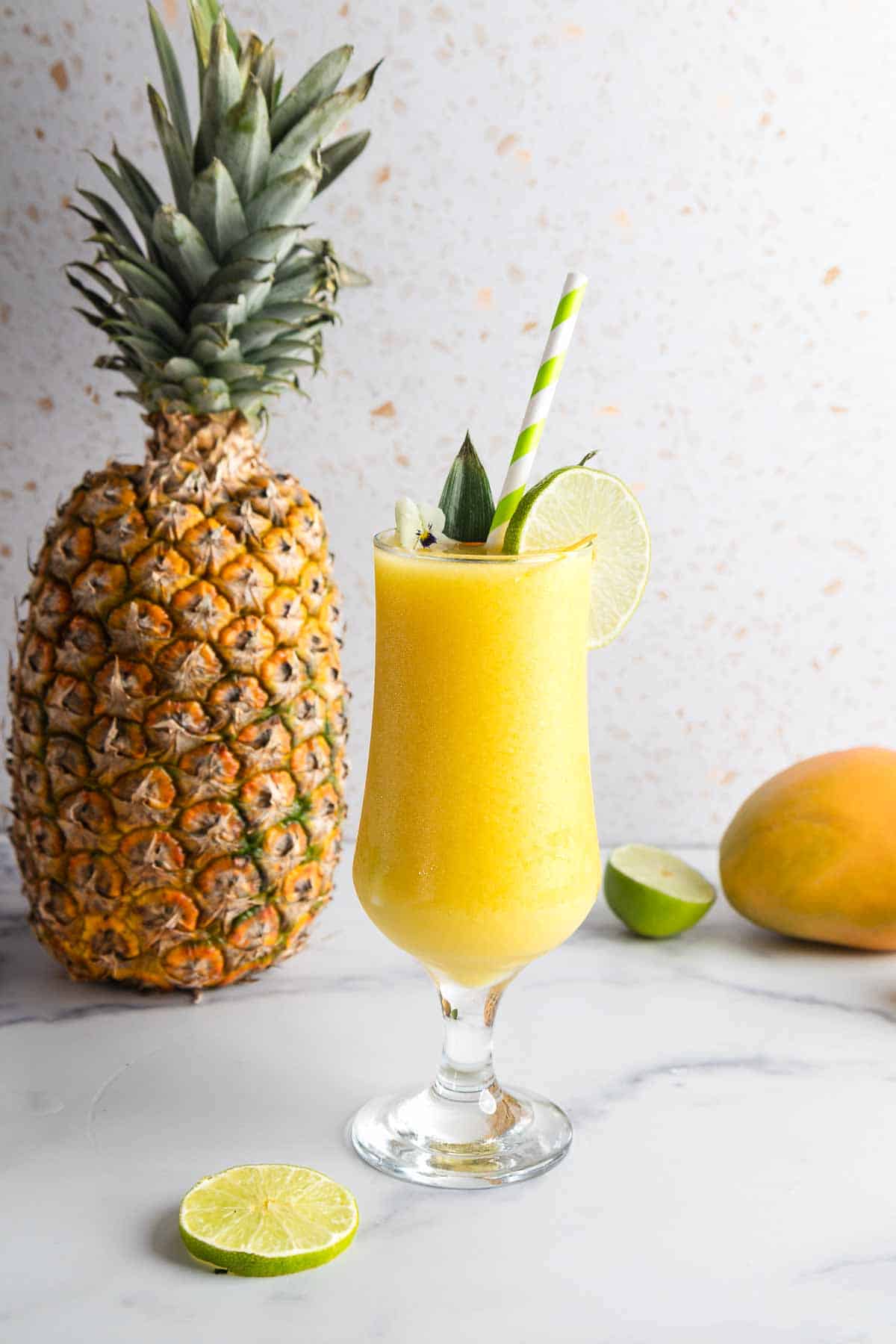A yellow tropical Mango Colada smoothie in a glass with a lime slice, next to a pineapple and mango on a marble surface.