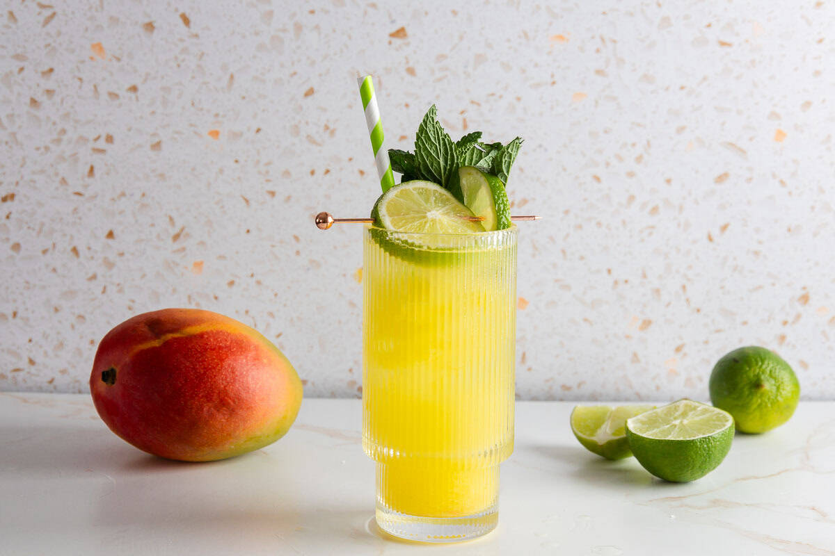 A yellow mango drink with ice, lime slices, and mint in a glass, next to a fresh mango and halved limes&mdash;perfect for a refreshing mocktail recipe.