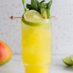 A yellow mango tropical drink with ice, mint, lime slices, and a striped straw in a glass on a white surface—a refreshing mocktail recipe perfect for summer.